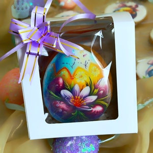 May include: A decorated Easter egg cookie in a clear plastic box with a purple ribbon. The cookie is decorated with a colorful floral design and a yellow background. The box is white with a clear window.