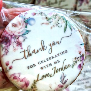May include: A round, white cookie with a floral design and the text "thank you for celebrating with me Love Jordan" in gold lettering.