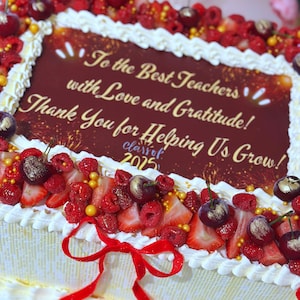 May include: A rectangular cake decorated with white frosting, fresh berries, and a red bow. The cake features a message in gold lettering: "To the Best Teachers with Love and Gratitude! Thank You for Helping Us Grow!" and "class of 2025".