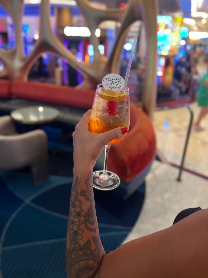 May include: A tall cocktail glass filled with an orange-colored drink, garnished with a cherry and orange slice. A white circular tag with the text "Thursday is little Friday" is attached to the glass. A clear straw is in the drink.