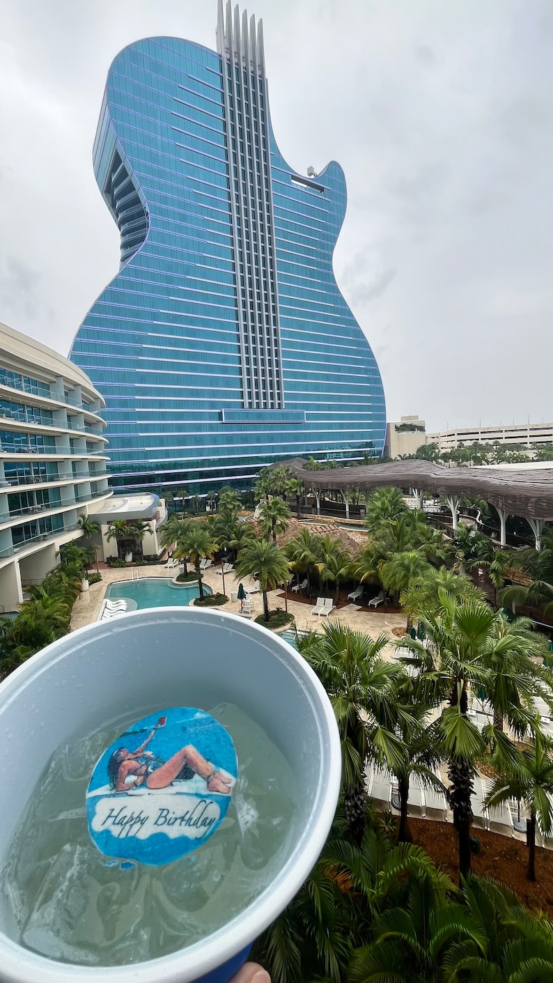 May include: A blue and white cup filled with ice and a decorative disc that says "Happy Birthday" with a woman on it. In the background is a guitar-shaped building and palm trees.