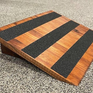 Peut inclure: Une planche inclinée carrée en bois avec une finition naturelle aux grains bruns. La surface présente trois bandes parallèles de ruban adhésif antidérapant noir disposées en diagonale, offrant une adhérence sur la plateforme en bois.