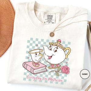May include: Ivory-colored t-shirt featuring a cartoon teapot and teacup design with a pink rose and a book. The design is set against a checkered background. The text "Everyday I'm Motherin'" is printed below the graphic.
