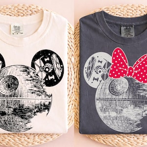 May include: Two t-shirts, one cream and one charcoal, each featuring a Death Star design with mouse ears and TIE fighters. The charcoal shirt has a red polka dot bow. The design is in black and white.
