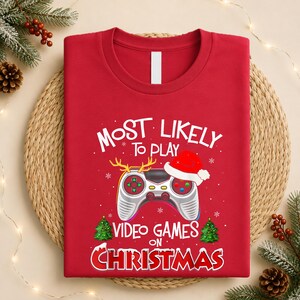 Op de afbeelding: Een rood T-shirt met ronde hals en een afbeelding van een videogamecontroller met een kerstmuts en rendiergewei. De tekst luidt: Most Likely To Play Video Games On Christmas, vergezeld door twee kleine groene kerstbomen als illustratie.