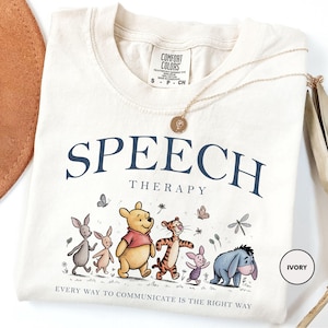 May include: An ivory t-shirt featuring the text "SPEECH THERAPY" and "EVERY WAY TO COMMUNICATE IS THE RIGHT WAY" with a graphic of Winnie the Pooh, Tigger, Piglet, Eeyore, and Roo. A gold-toned necklace with a circular pendant rests on the neckline.