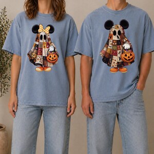 Peut inclure: Deux t-shirts bleus arborant un motif de fantôme portant des oreilles de souris et un drap à motifs patchwork. Le fantôme de gauche porte un nœud jaune à pois, et les deux tiennent un petit seau citrouille orange rempli de bonbons.