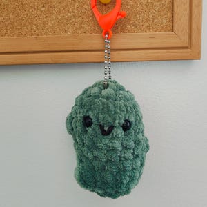 May include: A handmade green plush keychain in the shape of a smiling character. The keychain has black eyes and a black stitched mouth. It hangs from a silver chain and an orange clip.