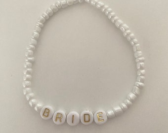 May include: White beaded bracelet with the word "BRIDE" spelled out in gold letters on white beads. The bracelet is made of small, round, white beads strung together. A simple and elegant accessory for a wedding.