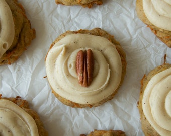 May include: Close-up of a batch of homemade carrot cake cookies with cream cheese frosting and pecan halves on top. The cookies are arranged on a white parchment paper background.