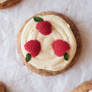 May include: Three raspberry topped cookies with white frosting and a sprig of mint on top. The cookies are on a white parchment paper background.
