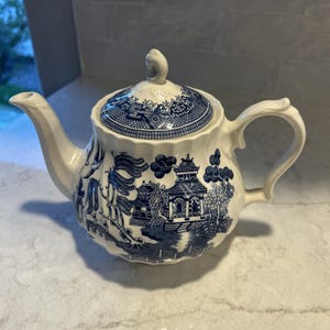 Op de afbeelding: Een crèmekleurige keramische theepot met een blauw willow-patroon met gedetailleerde illustraties van pagodes, bomen en bruggen. De theepot heeft een geschulpte behuizing, een gebogen handvat, een lange tuit en een bijpassend deksel met knop.