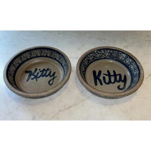 May include: Two round ceramic bowls with a speckled blue and tan rim. The interior of each bowl features a smooth tan base with the word Kitty written in dark blue script. The bowls have a low profile and a wide opening for easy access.