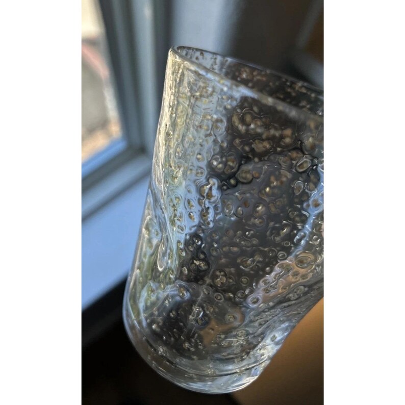 May include: A clear glass with a textured, bubble-like pattern. The glass has a cylindrical shape and appears to be partially filled. The light reflects off the surface, creating highlights and shadows. The glass is held in front of a window.