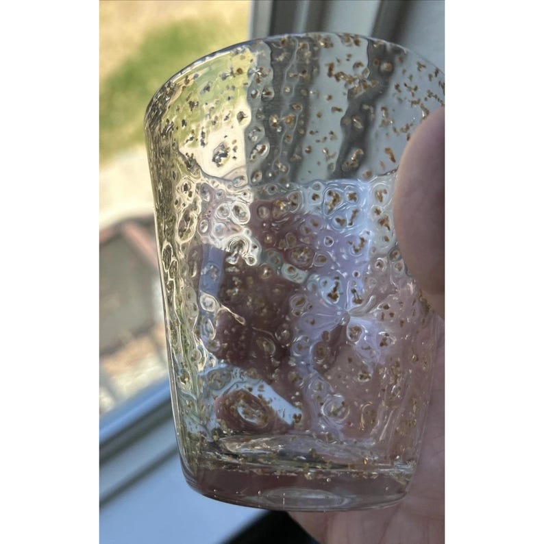 May include: A clear glass tumbler with a textured surface and gold flecks. The glass has a short, cylindrical shape and is held by a hand. The glass is partially filled with a clear liquid.