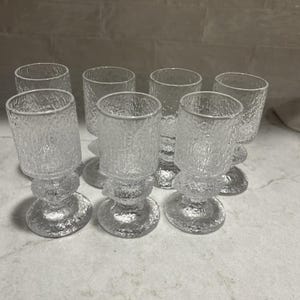 May include: A set of seven clear glass goblets featuring a textured, bark-like finish on the exterior. Each goblet has a cylindrical bowl, a thick stem with a rounded decorative node, and a circular base, all crafted from transparent glass.