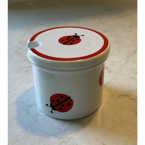 May include: White ceramic jar with a lid featuring a red rim and a red ladybird design. The jar has a small handle and additional ladybird illustrations on the side. A charming kitchenware item.