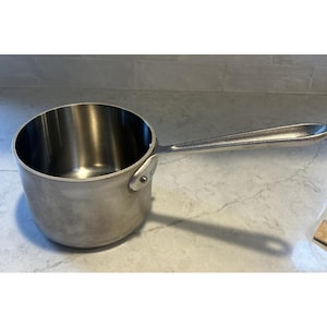 May include: A stainless steel saucepan with a long handle. The small cooking pot is silver and has a rounded body. The handle is riveted to the side of the pot. The saucepan is designed for cooking on a stovetop.