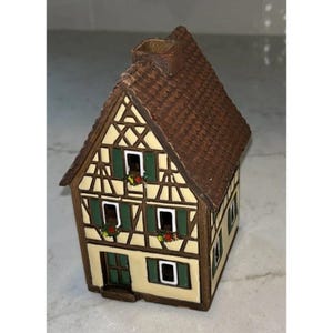 May include: A ceramic house-shaped candle holder featuring a brown textured roof with a chimney, cream-colored walls with dark brown timber framing, green shutters, and small painted flower boxes beneath the windows on the front facade.