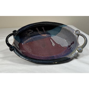 Puede incluir: Una fuente de cerámica ovalada con dos asas laterales, con un esmalte brillante en tonos negro profundo, morado oscuro y azul apagado. La superficie interior muestra un acabado mezclado y multicolor con bordes curvos y suaves.