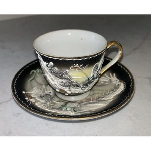 May include: A porcelain teacup and saucer set featuring a black and white gradient background with a raised, textured white dragon motif. The cup has a gold-toned handle and rim, while the saucer displays a matching dragon design with gold trim.