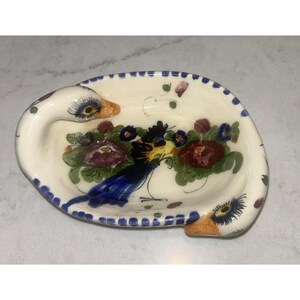 May include: An oval ceramic dish featuring two sculpted bird heads on opposite ends. The interior is decorated with a hand-painted floral motif in shades of purple, red, blue, and green. The rim is accented with a repeating pattern of blue dashes.