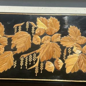 May include: A rectangular black box featuring a decorative inlay of golden straw leaves and catkins on the lid. The box has a thin gold border around the edges and small gold-colored circular accents at each of the four corners of the lid.