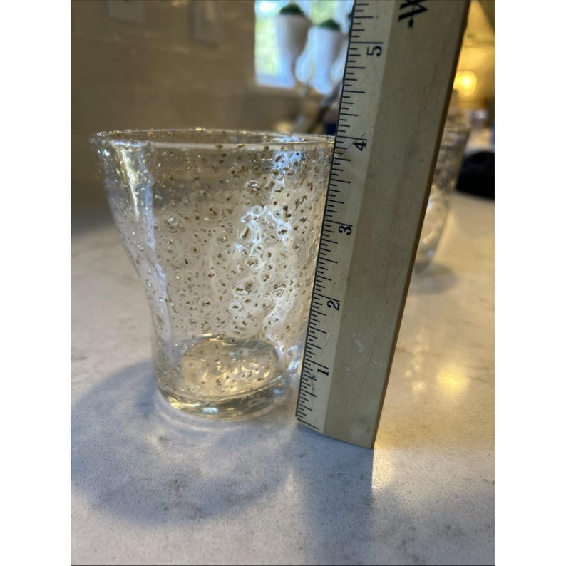 May include: A clear glass with a textured surface. The glass is approximately 3 inches tall, as measured by a wooden ruler. The glass has a unique, irregular shape and is suitable for beverages.