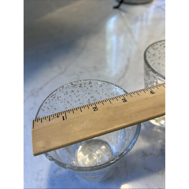 May include: Clear glass votive candle holders with a speckled design. A wooden ruler shows the diameter of the glass is approximately 3 inches. The votives are on a white surface.