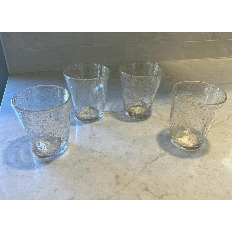May include: Four clear drinking glasses with a textured, speckled design. The glasses have a slightly curved shape and are arranged on a light-colored countertop. The glassware is suitable for water, juice, or other beverages.