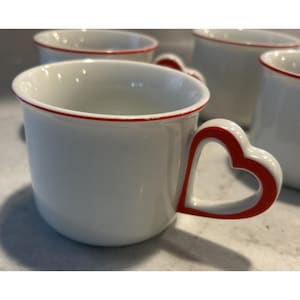 May include: White ceramic mugs with a red heart-shaped handle and a red rim. The mugs are grouped together, highlighting their design. The heart-shaped handle gives a unique touch to the classic mug shape.