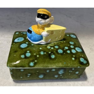 May include: A rectangular ceramic box with a glossy olive green glaze featuring light blue circular spots. The lid is topped with a sculpted white mouse figure wearing a yellow sweater, blue pants, and a black eye mask, holding a piece of yellow Swiss cheese.