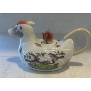 May include: A white ceramic teapot shaped like a duck with a brown head and beak. The body features painted illustrations of birds on branches, butterflies, and insects. A small orange flower-shaped knob sits on the lid, and a curved handle extends from the back.
