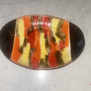 Op de afbeelding: Een ovale keramische schaal met een centraal paneel met verticale strepen in feloranje, lichtgeel en donkerbruin. Het centrale gedeelte heeft een getextureerd rasterpatroon, terwijl de twee uiteinden van de schaal effen donkerbruin zijn.