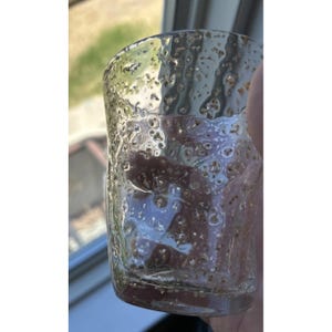 May include: A clear, textured glass with a bubbly surface. The glass has a cylindrical shape and is filled with small, golden-brown particles. The glass is held up against a window, with natural light illuminating the interior.