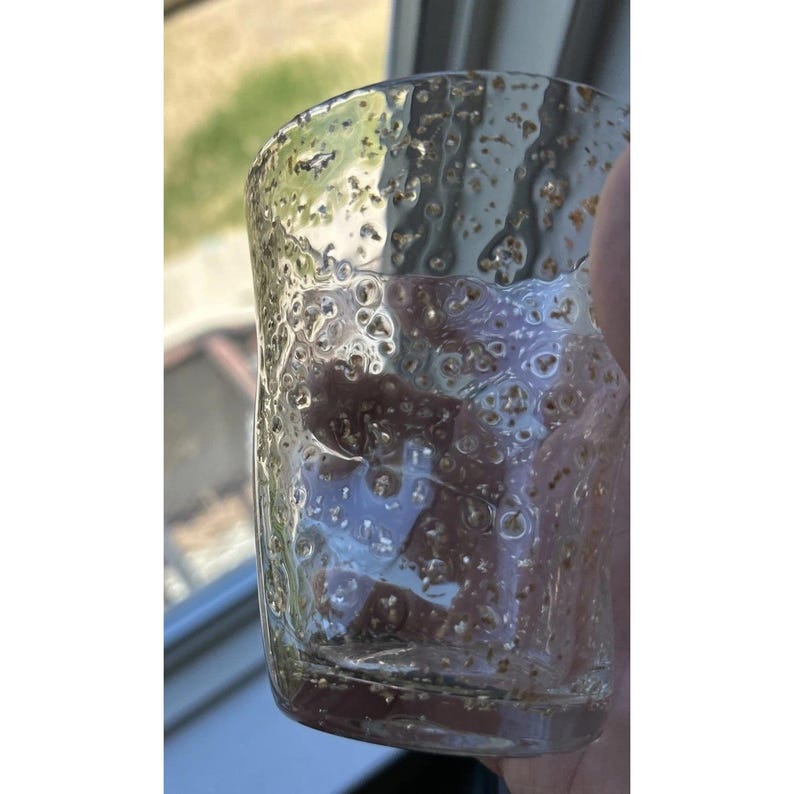 May include: A clear, textured glass with a bubbly surface. The glass has a cylindrical shape and is filled with small, golden-brown particles. The glass is held up against a window, with natural light illuminating the interior.