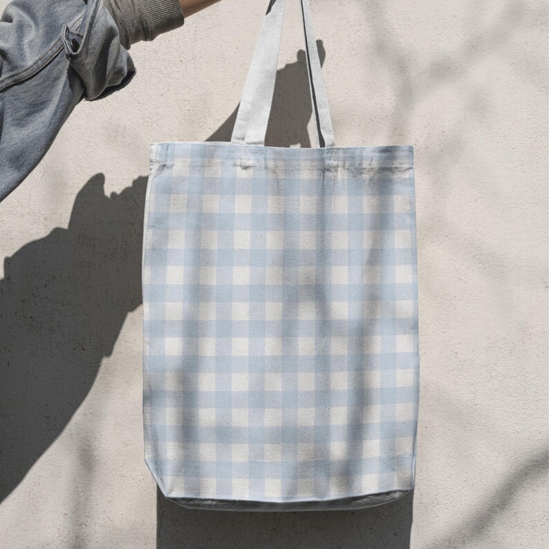 Puede incluir: Un bolso tote a cuadros azul claro y blanco con correas a juego se muestra. El patr&oacute;n del bolso es un dise&ntilde;o cl&aacute;sico de cuadros vichy, perfecto para llevar comestibles o art&iacute;culos personales. El bolso es de tela.