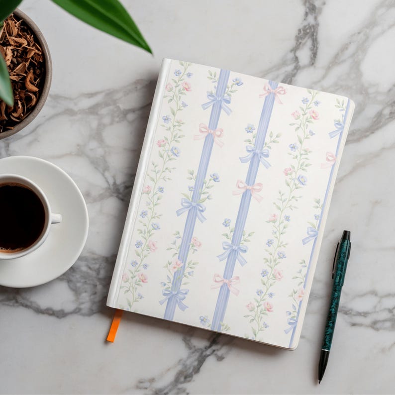 Puede incluir: Un cuaderno blanco con un dise&ntilde;o floral y de lazo, con detalles en azul y rosa. Una taza de caf&eacute;, un bol&iacute;grafo verde y una planta est&aacute;n sobre una superficie de m&aacute;rmol.
