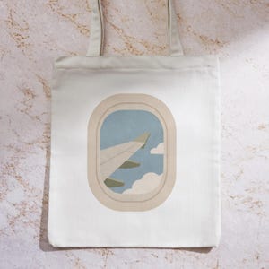 May include: White tote bag featuring an illustration of an airplane wing viewed through a window. The design includes a light blue sky, white clouds, and a beige window frame. The bag has two beige straps.