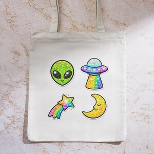 K&ouml;nnte beinhalten: Wei&szlig;er Canvas-Tote-Bag mit vier glitzernden Designs zum Thema Aliens. Enth&auml;lt einen gr&uuml;nen Alienkopf, ein UFO, einen Sternschnuppen und einen Mond. Die Designs sind schwarz umrandet und haben ein funkelndes Finish.
