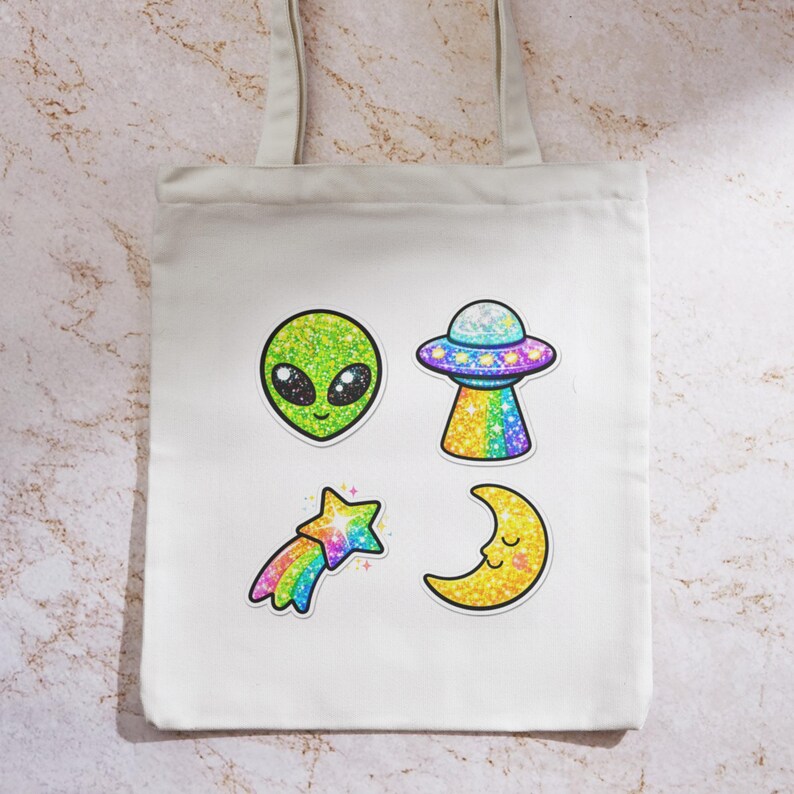 K&ouml;nnte beinhalten: Wei&szlig;er Canvas-Tote-Bag mit vier glitzernden Designs zum Thema Aliens. Enth&auml;lt einen gr&uuml;nen Alienkopf, ein UFO, einen Sternschnuppen und einen Mond. Die Designs sind schwarz umrandet und haben ein funkelndes Finish.
