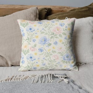 Puede incluir: Coj&iacute;n decorativo cuadrado con un estampado floral. El coj&iacute;n presenta un fondo color crema con un patr&oacute;n repetido de flores azules, rosas y amarillas con tallos y hojas verdes. El coj&iacute;n est&aacute; sobre una cama con otros cojines.