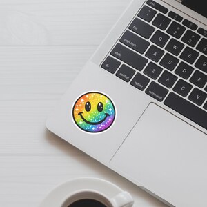 K&ouml;nnte beinhalten: Ein runder Aufkleber mit einem Regenbogen-Smiley-Gesicht, verziert mit Glitzer und Sternen. Das Smiley-Gesicht hat einen schwarzen Umriss und befindet sich auf wei&szlig;em Hintergrund. Der Aufkleber liegt auf einem Laptop.
