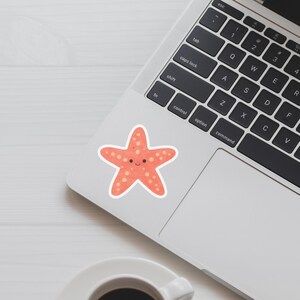 May include: A coral-colored starfish sticker with a smiling face and small, light-colored spots. The sticker is on a laptop, near the keyboard and a white coffee cup.