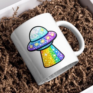 K&ouml;nnte beinhalten: Wei&szlig;e Keramiktasse mit einem bunten, glitzernden UFO-Design. Das UFO hat ein blaues, lila und gelbes Farbschema mit einem Regenbogenstrahl. Die Tasse steht in einer wei&szlig;en Box mit braunem Verpackungsmaterial.
