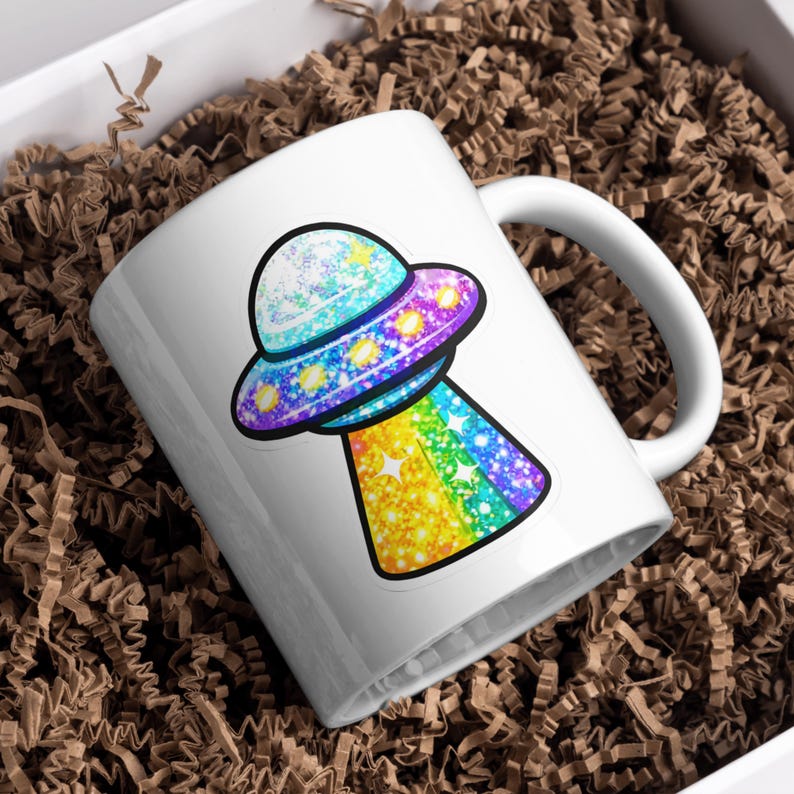 K&ouml;nnte beinhalten: Wei&szlig;e Keramiktasse mit einem bunten, glitzernden UFO-Design. Das UFO hat ein blaues, lila und gelbes Farbschema mit einem Regenbogenstrahl. Die Tasse steht in einer wei&szlig;en Box mit braunem Verpackungsmaterial.