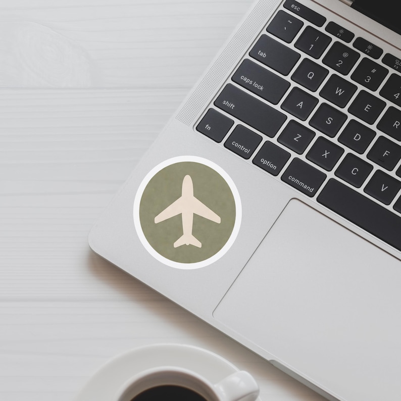 May include: A round sticker featuring a cream-colored airplane silhouette on a textured olive green background, encircled by a white border. The sticker is placed on a laptop keyboard.