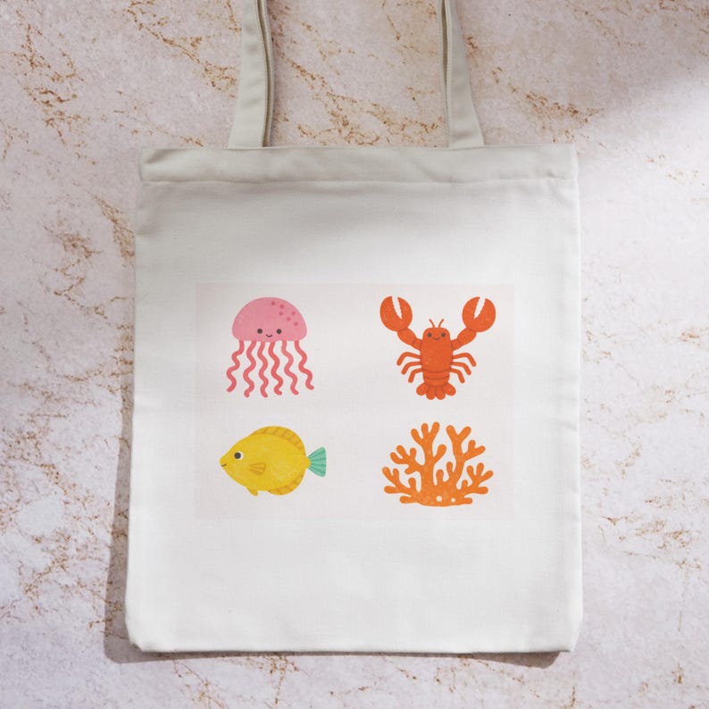 May include: A white tote bag featuring four colorful sea-themed illustrations: a pink jellyfish, an orange lobster, a yellow fish, and orange coral. The bag has two handles and is made of a light-colored fabric.