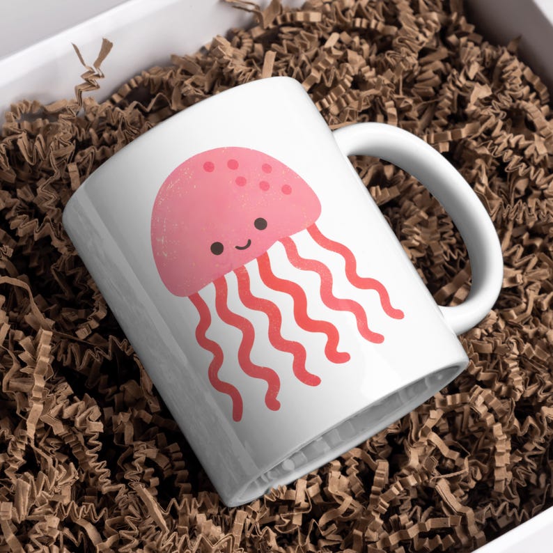 May include: White ceramic mug featuring a pink cartoon jellyfish with a smiling face and wavy red tentacles. The mug has a white handle and is set against a background of brown packing material.