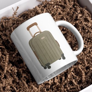 May include: White ceramic mug featuring a green suitcase illustration with brown accents. The suitcase has a retractable handle and wheels. The mug is set against a background of brown packing material.